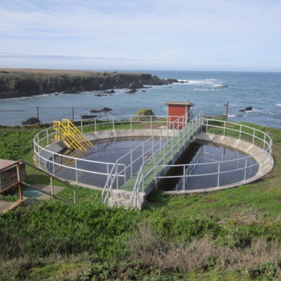 An overhead view of an open-air round clarifying tank at a wastewater treatment facility on a clear day. With the opening at ground level, the tank is surrounded by handrails and bisected by a catwalk that leads to a small red shed. The tank is located in a grassy elevated coastal area overlooking the ocean, with a cliff visible in the back left.
