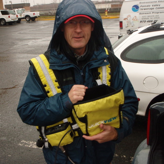 An inspector stands in a large parking lot in a light rain. He is holding a fiber optic camera and wearing a yellow high-visibility vest over a blue jacket with the hood up. A few white work vehicles are parked in the background.