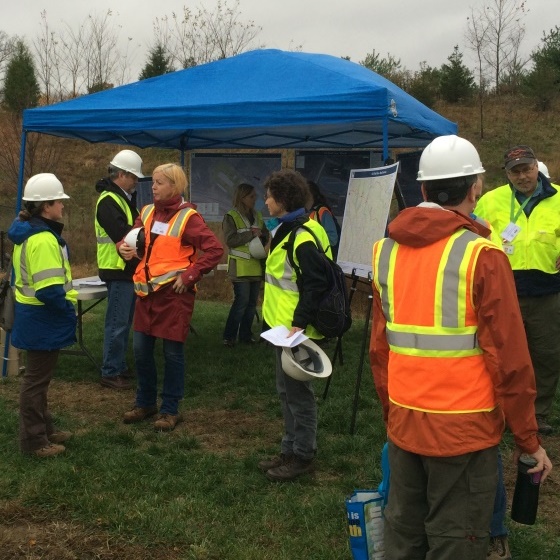 People wearing high visibility safety vests and hard hats gathered around a blue tent shelter on an overcast day. The area is grassy and lightly forested, with pine trees in the distance.