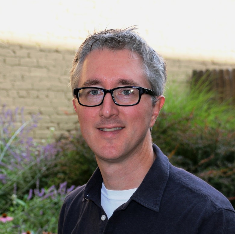 James Ashby, IT and GIS Team Leader, stands outside wearing business casual attire in front of a flowering garden area and a sunlit light-colored brick background.