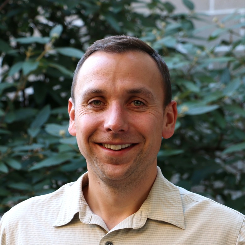 Kort Kirkeby, Team Leader, Ecological Services, stands outside wearing business casual attire in front of a leafy green background.