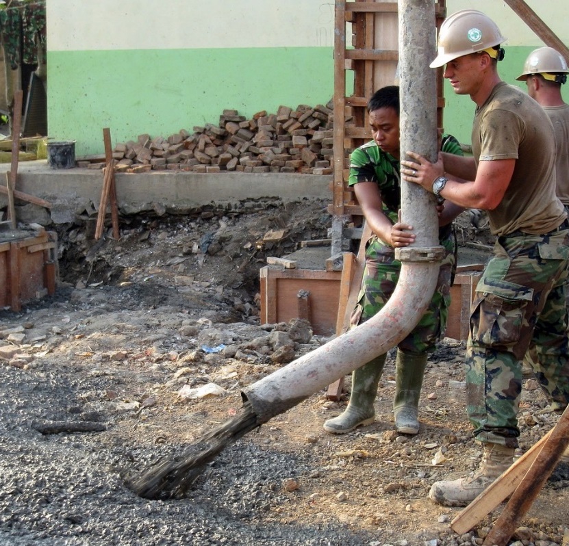 People dressed in green camouflage with hard hats are building a foundation, directing the flow of concrete out of a grey pumping hose hanging from above. A pile of bricks is visible in the background in front of a white and green exterior wall.