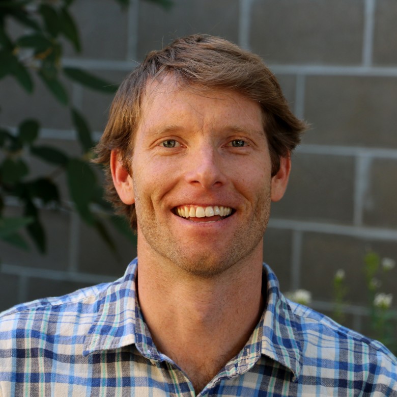 Bobby Jacobsen, Colorado Operations Manager and Environmental Scientist, stands outside wearing business casual attire in front of a light-colored brick wall and some leafy plants.