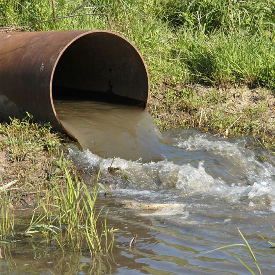 A large brown steel drainage pipe discharging into a stream surrounded by grasses. Light brown water is rippling out of the pipe and meeting the receiving water.