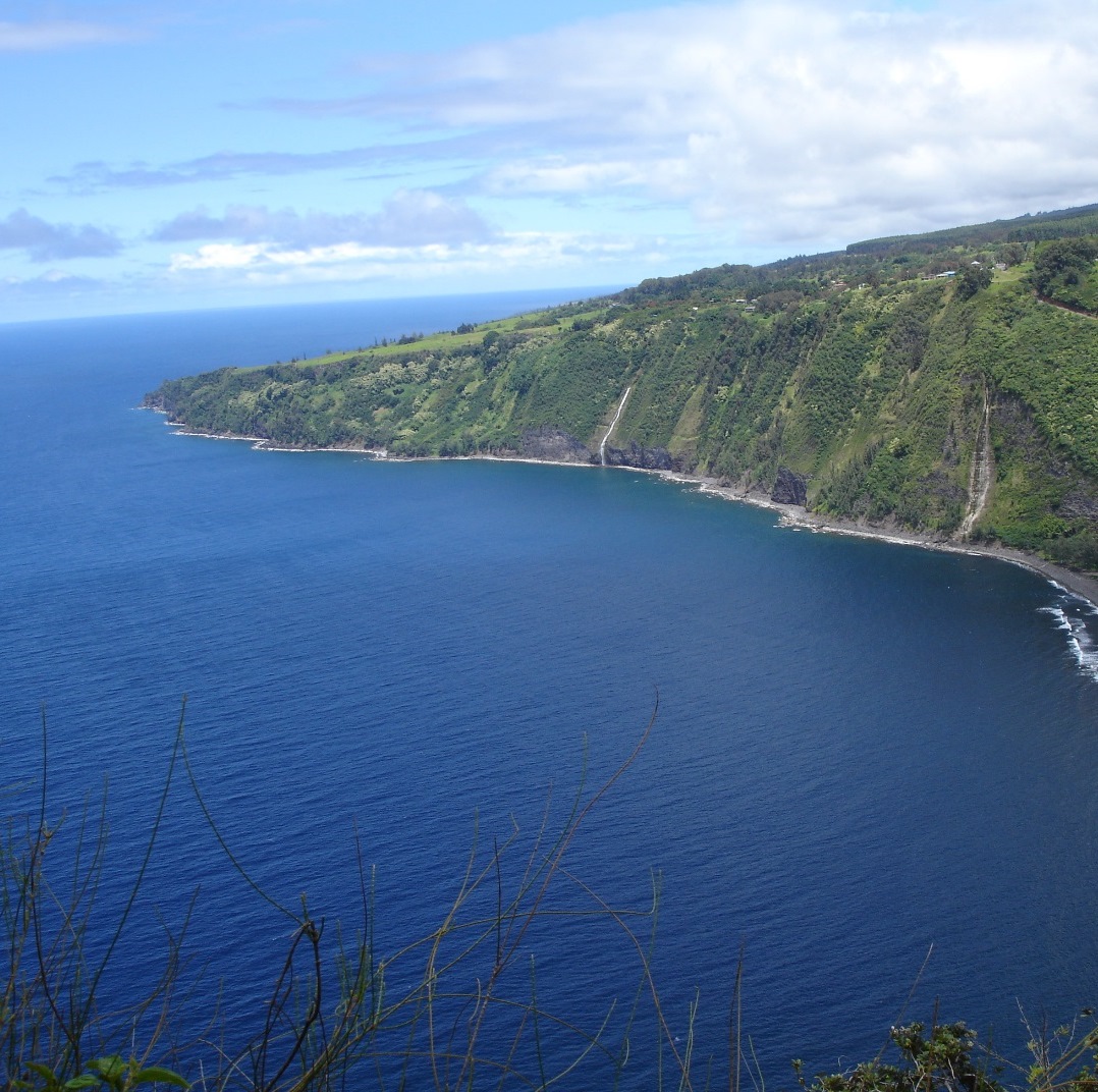 View looking down towards the deep blue ocean on a calm, partly cloudy day. A section of lush green coastline sticks out into the ocean from the upper right side of the photo.