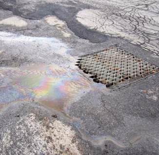 Looking down at a storm drain in a parking lot. It is surrounded by sand and a rainbow-colored oil sheen flowing into it.