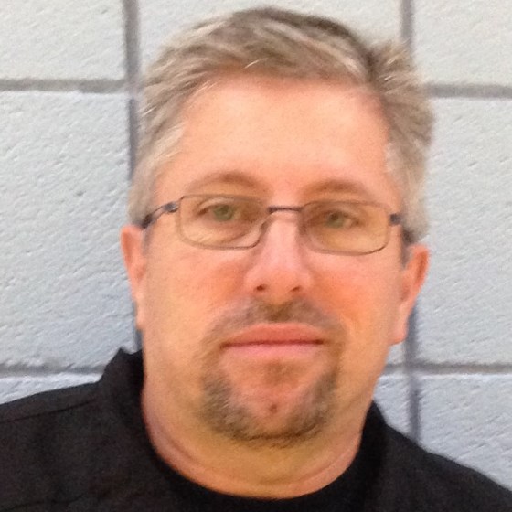 Chuck Durham, Principal Engineer and Senior Project Manager, stands in business casual attire in front of a light-colored brick background.