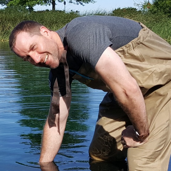 Dan Connally, Virginia Operations Manager and Senior Permit Writer, wades in a stream up to his knees. He is bent over with his hand in the still water, wearing chest waders and a grey tee shirt. A grassy stream bank is visible in the background.