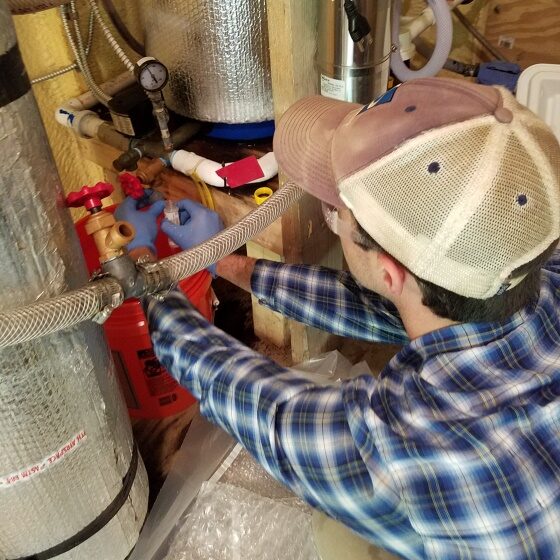 A person wearing a hat, blue and white plaid shift, and blue rubber gloves works on well plumbing.