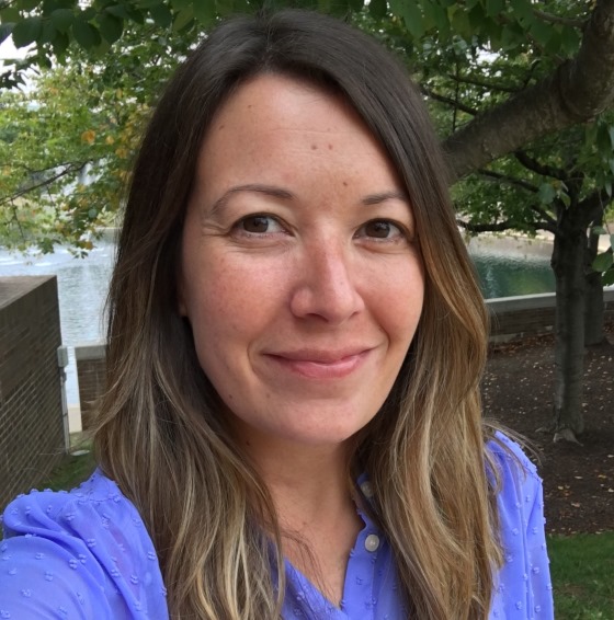 Hilary Rains, Environmental Scientist, stands outside in business casual attire in front of leafy green trees and a pond in the background.