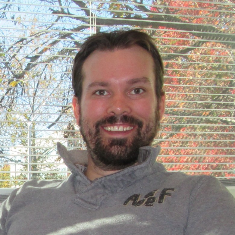 Jake Albright, Environmental Scientist, sits in front of a window with open blinds wearing business casual attire. Trees are showing their fall foliage in the background.