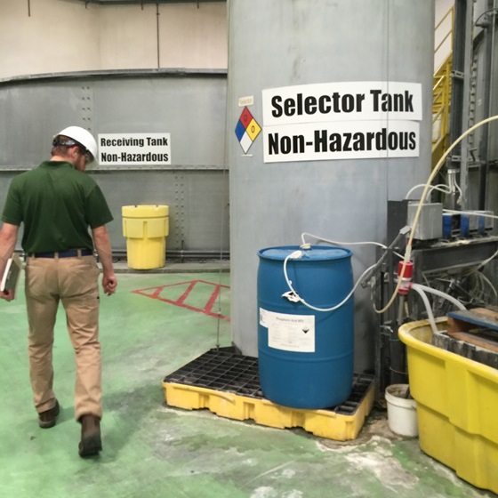 An inspector walking inside a building wearing a white hardhat and looking down at a chemical containment area beneath a fifty-gallon drum. The drum is sitting in front of a grey tank marked “Selector Tank Non-Hazardous.” The green floor has white residue around the yellow plastic containment area.