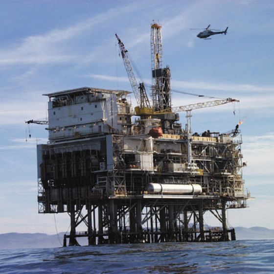 An offshore oil rig stands above the ocean surface while a helicopter flies above and to the right. It is a clear, sunny day with a faint mountainous coastline visible in the distance.