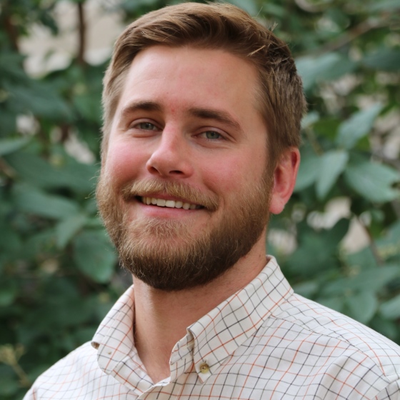 Stephen Clark, Environmental Scientist, stands wearing business casual attire in front of a leafy green background.