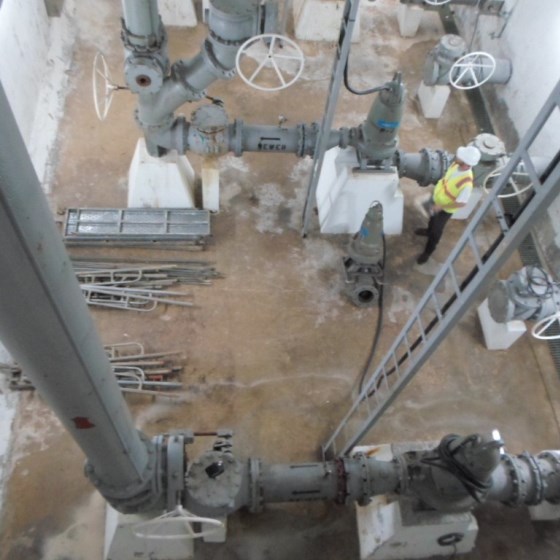 An overhead view of a pipe galley in a concrete pit with grey piping, valves, and pumps. A spare pump sits on the floor next to a person in a yellow safety vest and hardhat preparing to replace it. Some grey scaffolding is disassembled on the floor to the left.