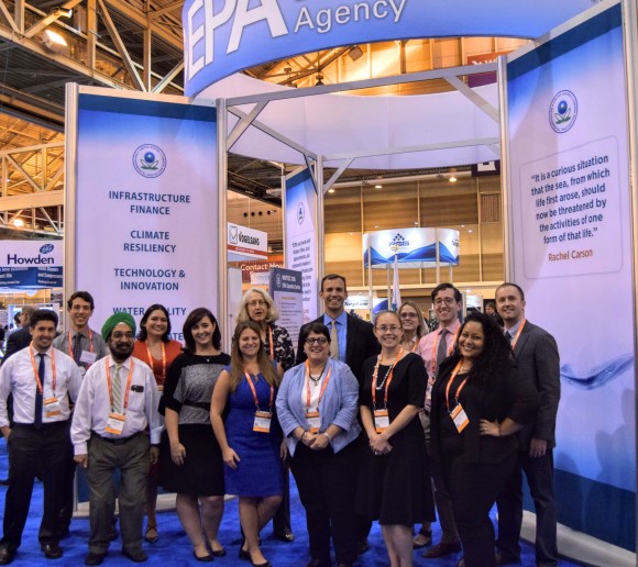A crowd of people dressed in business attire with lanyards around their necks gather together in the foreground to pose for a photo. They are standing in front of EPA’s tall exhibit booth, featuring blue and white informational signage inside a convention hall.