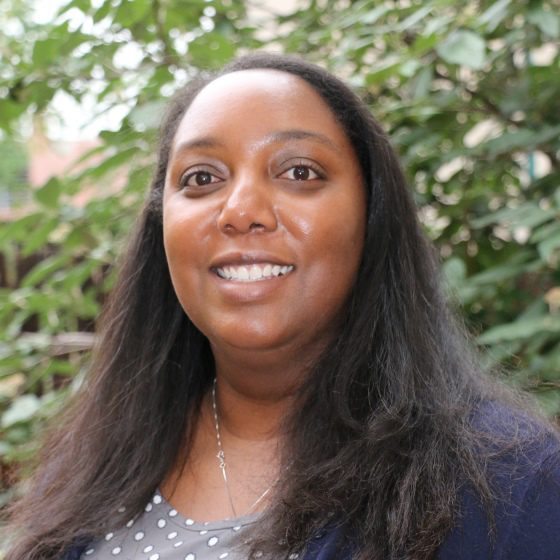 Yatasha Moore, Environmental Engineer, stands outside wearing business casual attire in front of a sunlit leafy green background.