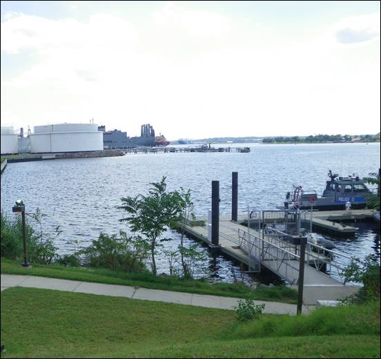 A view of Chesapeake Bay standing from shore and looking out. A grassy area in the foreground includes a walkway going out to a boat dock. The silvery water of the bay stretches off into the distance, while large white tanks and an industrial area line the left banks.
