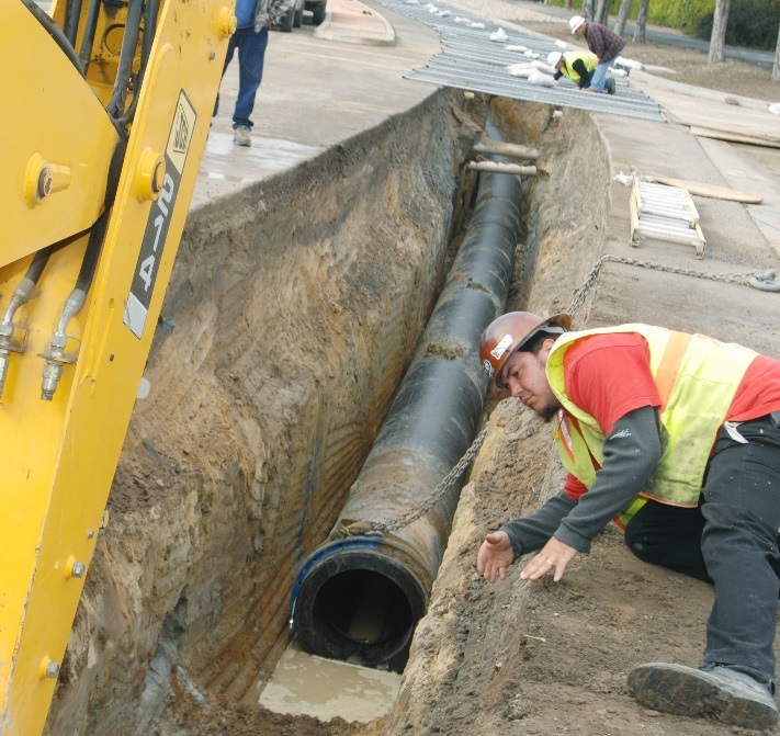 A person in a yellow safety vest and hard hat lays down on the right, watching an excavator dig a trench for a water pipe. On the left the large, yellow excavator arm extends down into the trench. In the distance, the rest of the work crew is connecting pipe as the trench gets opened and graded.