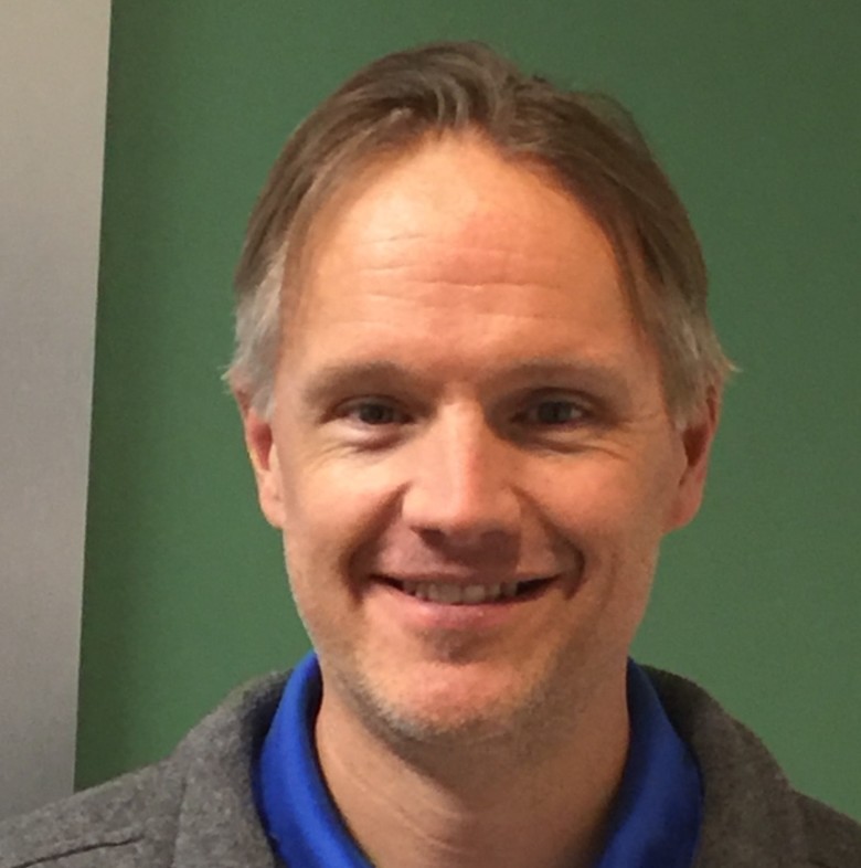 Jason Rose, Senior Environmental Engineer, stands in business casual attire in front of a white and green wall.