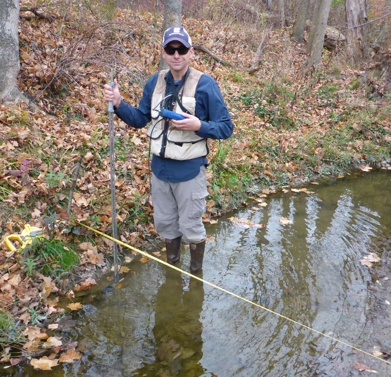An environmental field technician wading in a shallow, brown stream holding a flow measuring device, wearing a life jacket, hat, and sunglasses. A measuring tape is stretched across the stream, which flows through a forested area with leaves on the ground.