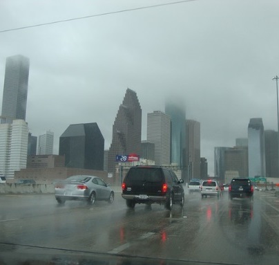 Looking out of a windshield, driving on a busy highway towards the city in a rainstorm. High-rise buildings line the horizon as they stick up and disappear into the clouds. All the other vehicles have their headlights on to see through the daytime fog and rain.
