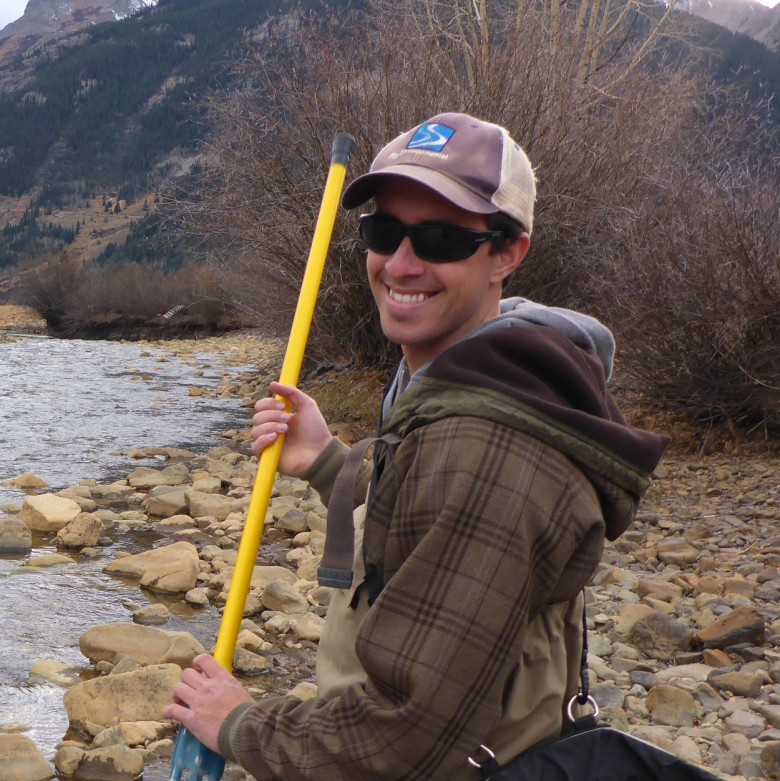 Zak Maurer-Erickson, Environmental Scientist, holding yellow field equipment and wearing a hat, jacket, and waders. A stream flowing from a pine tree covered mountain is visible in the background.