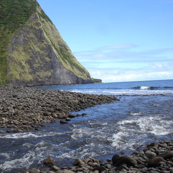 Standing at a stream flowing into the ocean on the coast of a Hawaiian island. A rocky coastline contrasts with the blue of the sky and ocean. A large, steep cliff meets the ocean in the background on the left.