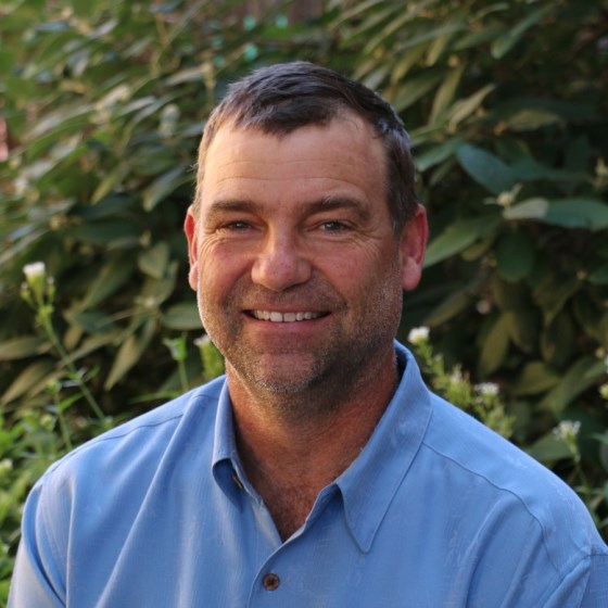 Wesley Ganter, Founder and CEO of PG Environmental, stands in business casual attire in front of a sunlit leafy green background.