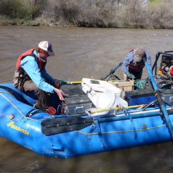 Standing on the bank next to a gear packed raft. Two people prepare to take samples on a free-flowing section a light brown colored river.