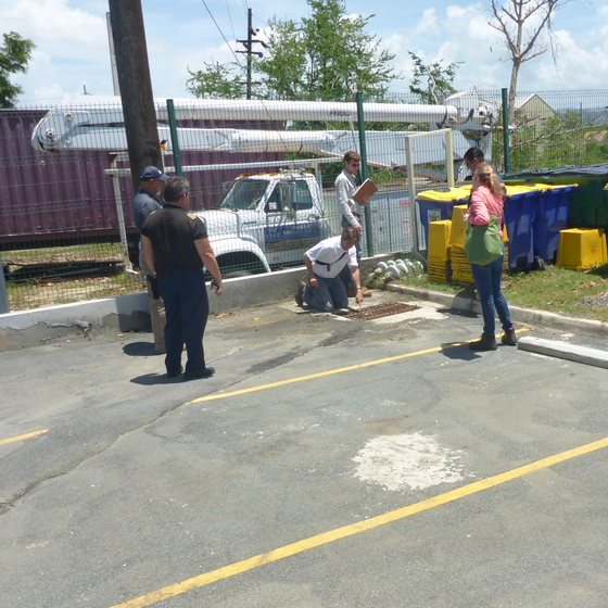 A group of people standing in a parking lot on a sunny day. The group is congregated around a storm drain, with one person pointing down to it. A service truck is parked in the background.