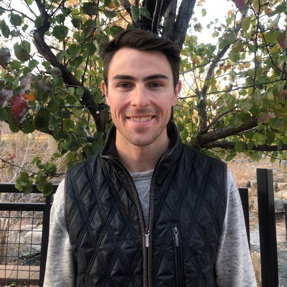 Alex Martinez, Environmental Scientist, stands outside wearing business casual attire with a sunlit tree and clear creek flowing in the background.