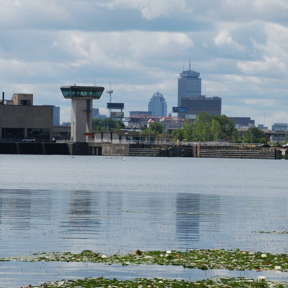 A skyline made up of buildings in the distance with a control tower standing at the edge of a lake. The sky is cloudy, but the water is calm.