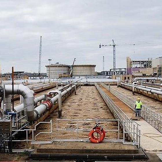 A man in a yellow safety shirt cleaning a walkway at a wastewater plant aeration basin. Several long pipes line the edges of long brownish basins. A view of a cloudy day looking towards a horizon with big tanks and cranes.