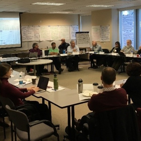 Multiple people sitting in a meeting room in a circle, water bottles and laptops on the tables and handwritten notes lining the walls.