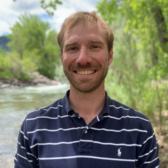 James Hollibaugh, Engineer, stands in business casual attire with a stream and trees in the background.