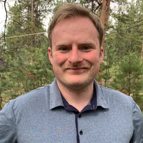 Andy Rimelman, Junior Engineer, stands outside in business casual attire in front of a dense pine forest in the background.