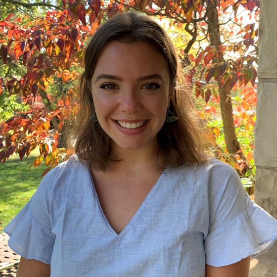 Emily Isaacs, Environmental Scientist, stands outside wearing business casual attire. Trees are showing their fall foliage in the background.