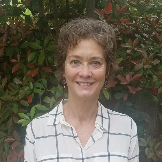 Lynn Kurth, Principal Scientist, stands outside wearing business attire in front of a green and red leafy background.