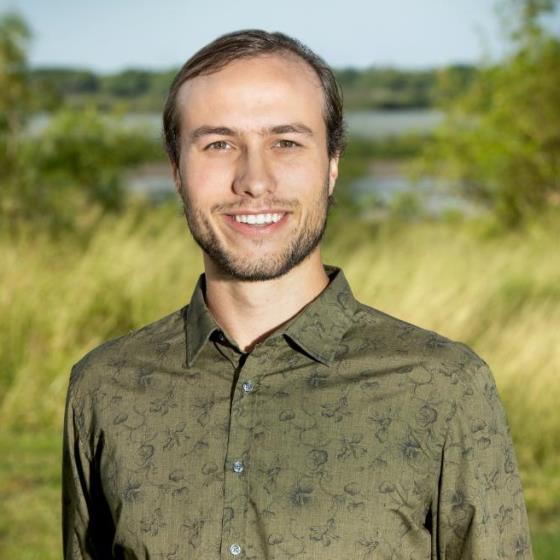 Abe Margo, Environmental Scientist, stands wearing business casual attire in front of a grassy landscape with a water body visible in the distance.