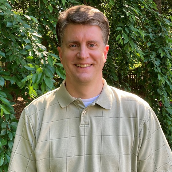 Kelly Meadows, Senior Environmental Scientist, stands wearing business casual attire in front of a leafy green background.