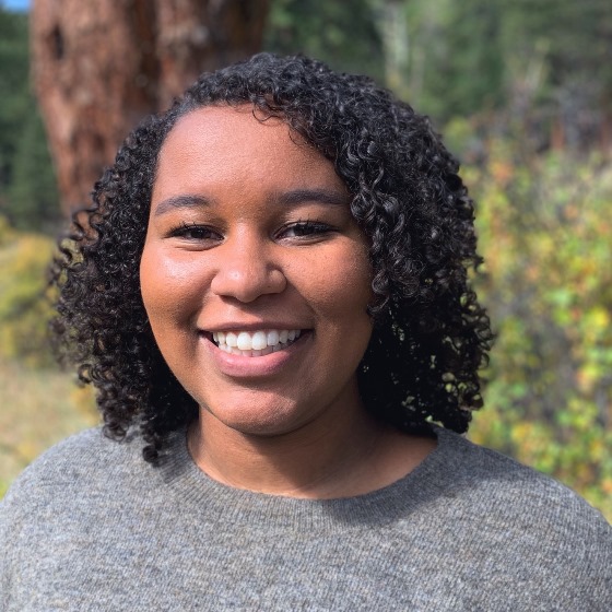 Sonrisa Macharia, Junior Engineer, stands wearing business casual attire in front of a leafy green background.