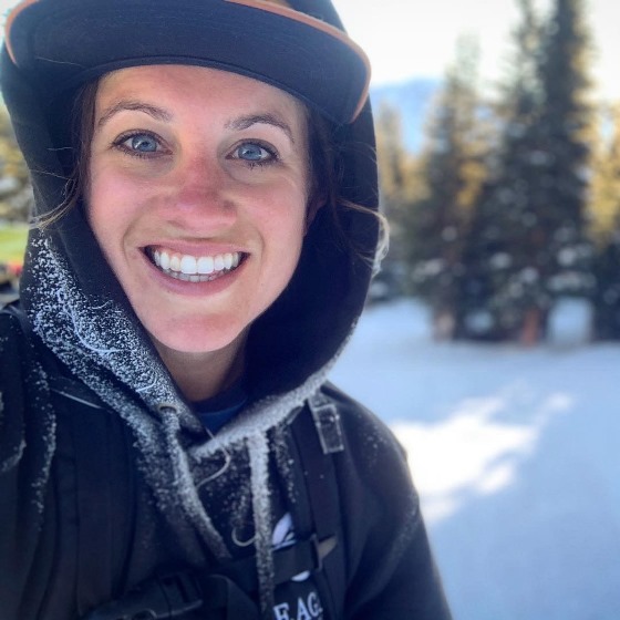 Kate Forsmark, Environmental Scientist, stands outside in casual attire in front of a snowy, forested landscape.