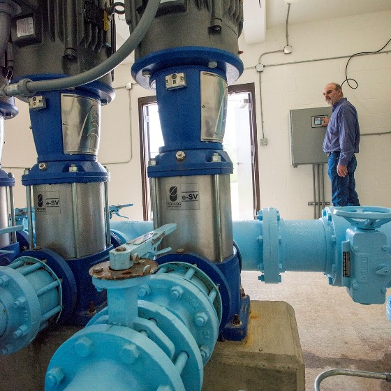 Blue and silver pipes at a water pumping station are pictured in the foreground as an operator makes adjustments through a square gray interface affixed to the far wall.