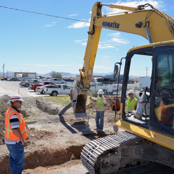 A yellow backhoe is parked at an active worksite next to a trench in the ground where new water lines will be laid. Three men in safety vests and hardhats stand nearby with a parking lot in the background and a blue sky overhead.