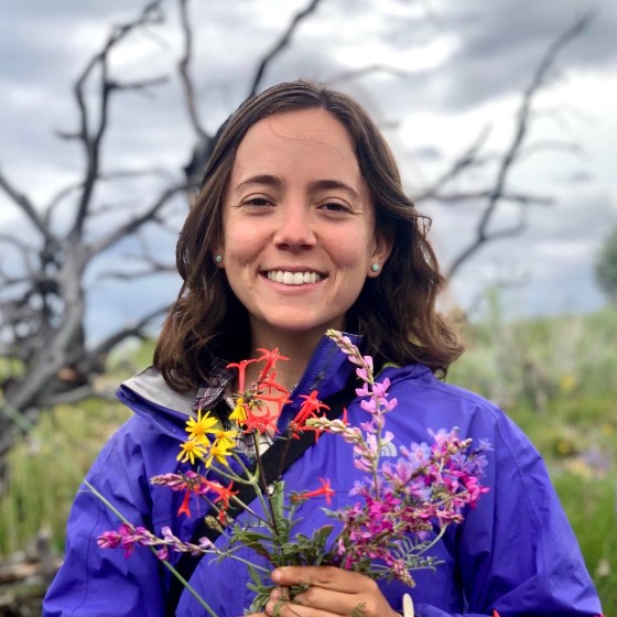 Megan Annis, Environmental Scientist, stands outside wearing business casual attire in a grassy landscape holding a colorful bouquet of wildflowers.