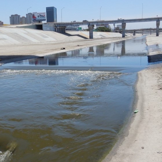 Water flows through a concrete conveyance in an urban setting spanned by a bridge in the background with skyscrapers visible in the distance.