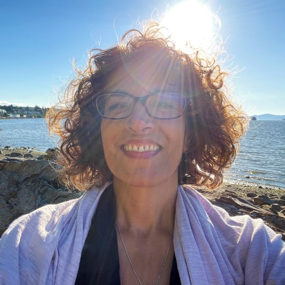 Tammy Cohen, Senior Environmental Scientist, stands in business casual attire on a rocky beach with a water body in the background and a bright sun in the cloudless sky.