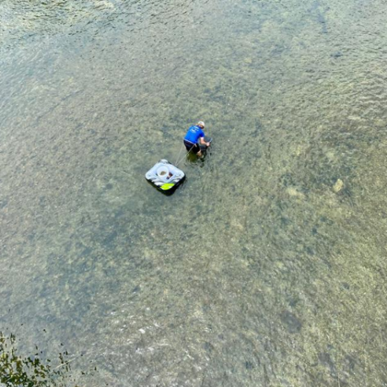 An aerial shot of a person in a blue shirt bending over in a shallow waterbody with a floatation device for sample collection tethered to his waist.