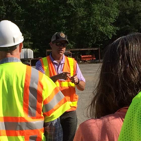 An instructor stands in a paved lot surrounded by green trees and faced by several people. Most wear high-visibility clothing, and one of the participants is also wearing a white hardhat.